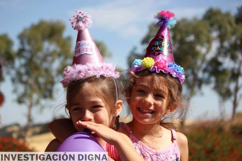 girl in left blowing purple balloon beside girl wearing pink birthday hat